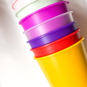May include: A stack of six colorful plastic cups, starting with a pink cup at the top and ending with a yellow cup at the bottom. The cups are stacked in a pyramid shape.