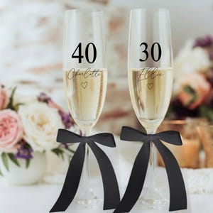 May include: Two champagne flutes filled with a bubbly drink. One glass has the number 40 and the name "Charlotte" with a heart, the other has 30 and "Ellie" with a heart. Both glasses are decorated with black bows.