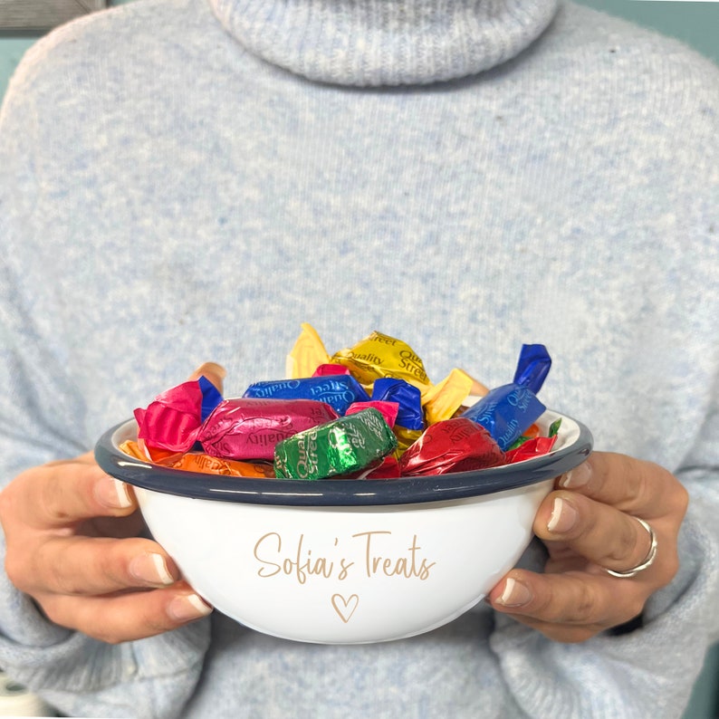 May include: White enamel bowl with a navy blue rim filled with colourful wrapped sweets. The bowl is personalised with the text "Sofia's Treats" and a heart symbol. The bowl is held by a person wearing a light blue jumper.