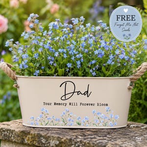 May include: A cream metal planter with rope handles, filled with blue forget-me-nots. The planter has the word "Dad" in script and the phrase "Your Memory Will Forever Bloom". A blue circle with text "FREE Forget Me Not Seeds" is in the top right.
