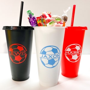 May include: Three black, white, and red reusable cups with straws. Each cup has a soccer ball design and a name: Lewis, Jaxon, and Harry.