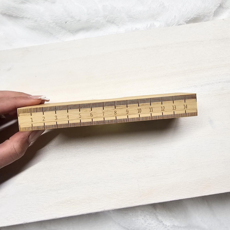 Puede incluir: Una regla de madera con marcas en pulgadas de 1 a 14. La regla es de madera de color claro y tiene marcas de color marr&oacute;n oscuro. La regla est&aacute; sujeta por una mano sobre un fondo blanco.