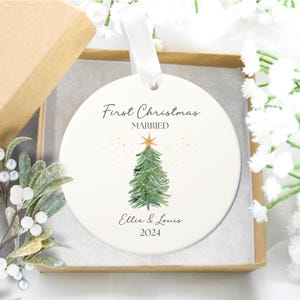 Puede incluir: Adorno navideño de cerámica blanca con una ilustración de acuarela de un árbol de Navidad con luces y una estrella en la parte superior. El adorno dice "First Christmas Married Ellie & Louis 2024".