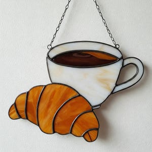 May include: Stained glass artwork depicting a white coffee cup with brown liquid and a golden-brown croissant. The piece is suspended by a chain and is suitable for kitchen or cafe decor.