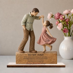 May include: A figurine of a person in a light green shirt and brown pants dancing with a child in a pink dress. The base features the text: TO MY DAD, Thank you for always being my safe place, my biggest supporter, and my guiding light.