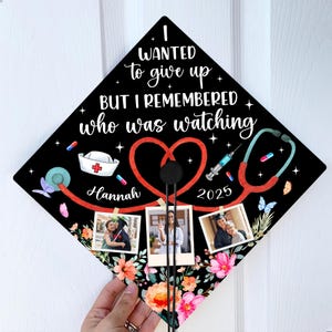 May include: Black graduation cap with a red heart, stethoscope, and the text "I wanted to give up but I remembered who was watching". The cap also has a nurse's hat, a syringe, and pills. The year 2025 and the name "Hannah" are written on the cap. Three photos are attached to the cap.