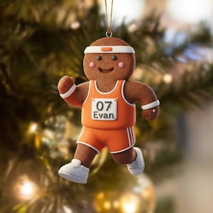 Può includere: Un ornamento per l'albero di Natale a forma di omino di pan di zenzero in posa da corsa, che indossa un'uniforme atletica arancione con dettagli bianchi. L'ornamento ha una fascia e scarpe bianche. Il numero "07" e il nome "Evan" sono sull'uniforme.