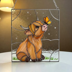 May include: A square stained glass panel showing a Highland cow and a butterfly. The cow is brown and orange, with curved horns, and is sitting in a field of green grass. A yellow and orange butterfly rests on the cow's nose. The panel has a silver metal frame.