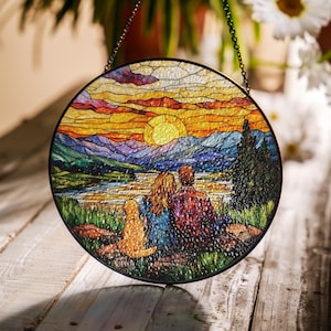 May include: A stained glass window hanging features a couple and a dog sitting on a grassy hill overlooking a lake with a sunset in the background. The mountains are in the distance and the sky is a vibrant orange and yellow.