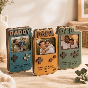 May include: Three wooden photo frames shaped like retro game consoles. Each frame has a different color and the words "DADDY", "PAPA", and "DAD" at the top. The frames contain photos of fathers and children. The text "THANKS FOR ALWAYS LEVELING US UP" is at the bottom.