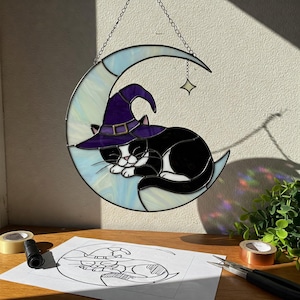 May include: Stained glass art piece featuring a black and white cat wearing a purple witch hat, sleeping on a crescent moon. The moon is light blue with a star hanging from a chain. A drawing of the cat and moon is on the table.