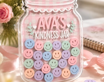 Könnte beinhalten: Ein durchsichtiger, glasförmiger Aufsteller aus Acryl mit einem rosa-weiß karierten Hintergrund. Der Text "AVA'S KINDNESS JAR" ist in Weiß aufgedruckt. Darin befinden sich zahlreiche kleine, bunte, runde Plättchen mit eingeprägtem Smiley.