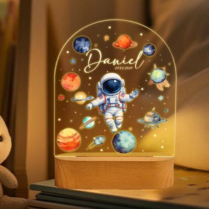 May include: A night light with a clear acrylic dome featuring an astronaut, planets, and stars. The base is made of wood and has the name "Daniel" and the date "27.02.2024" printed on it.