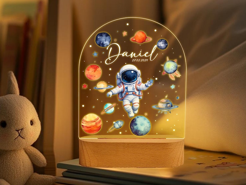 May include: A night light with a clear acrylic dome featuring an astronaut, planets, and stars. The base is made of wood and has the name "Daniel" and the date "27.02.2024" printed on it.