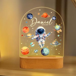 May include: A night light with a wooden base and a clear acrylic panel featuring an astronaut, planets, and stars. The panel is illuminated from behind, creating a soft glow. The text "Daniel 27.02.2021" is printed on the panel.