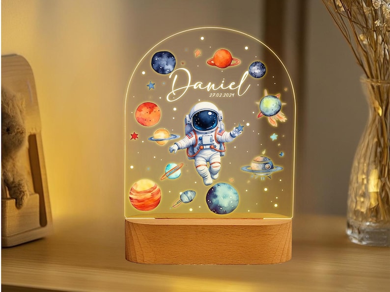 May include: A night light with a wooden base and a clear acrylic panel featuring an astronaut, planets, and stars. The panel is illuminated from behind, creating a soft glow. The text "Daniel 27.02.2021" is printed on the panel.