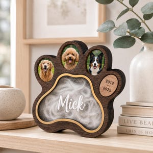 Op de afbeelding: Een donkerbruin houten pootvormig gedenkkader voor huisdieren met drie ovale fotovakken met afbeeldingen van honden. Het middengedeelte toont de naam Mick in wit schrift over witte vacht, met de jaren 2012-2025 gegraveerd op een ovaal.