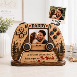 Op de afbeelding: Een houten fotolijst in de vorm van een gamecontroller. Het heeft een foto van een vader en kind, met de woorden "DADDY" en "My Girl". De lijst heeft ook de tekst "To the world, you are a Dad, but to us you are The World."