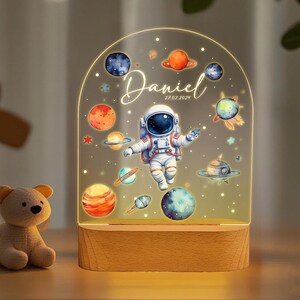 May include: A wooden night light with a clear acrylic panel featuring an astronaut, planets, and stars. The panel is illuminated from behind, creating a soft glow. The text "Daniel 27.02.2021" is printed on the panel.