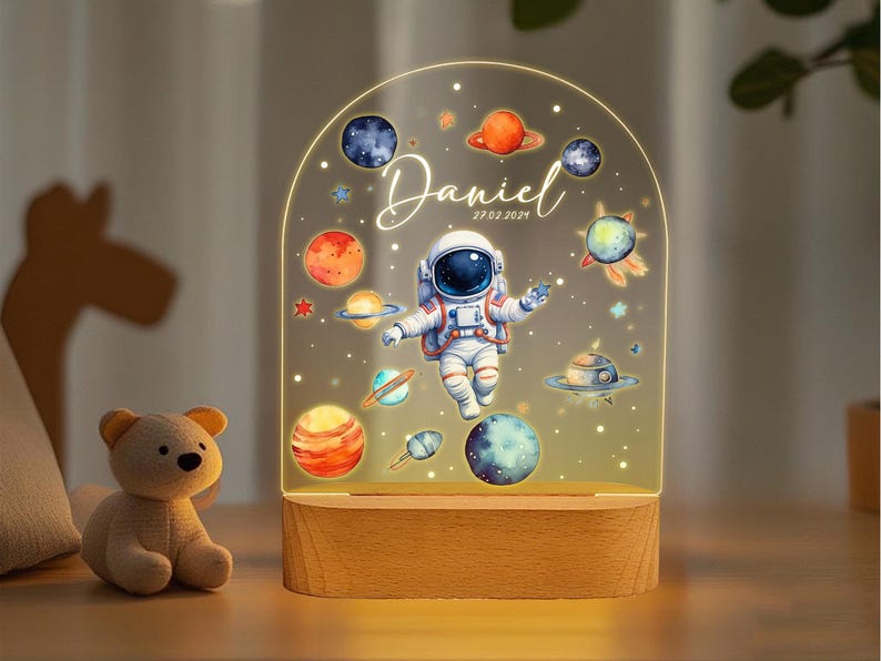 May include: A wooden night light with a clear acrylic panel featuring an astronaut, planets, and stars. The panel is illuminated from behind, creating a soft glow. The text "Daniel 27.02.2021" is printed on the panel.