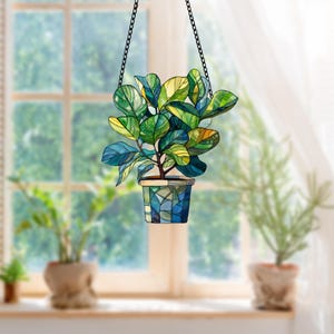 May include: A stained glass hanging decoration of a potted plant with green leaves and a blue and green mosaic pot. The plant is hanging from a black chain.