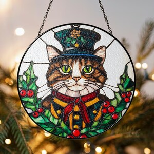 Op de afbeelding: Een hangend ornament van gekleurd glas voor het raam met een kat die een groen-zwarte hoed draagt met een rode sjaal en hulstbladeren rond de randen.