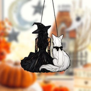 以下が含まれることがあります： 尖った帽子をかぶった黒い魔女と、黒いリボンをつけた白い猫が描かれたステンドグラスの窓飾り。魔女と猫は、鑑賞者から背を向けている。
