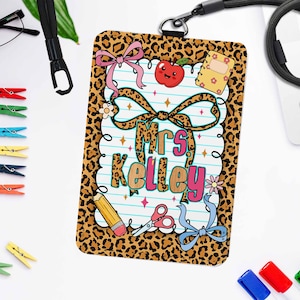 May include: A teacher's ID badge with a leopard print border and the name "Mrs. Kelley" in the centre. The badge features illustrations of an apple, notebook, pencil, scissors, and bows. A black lanyard is attached to the badge.