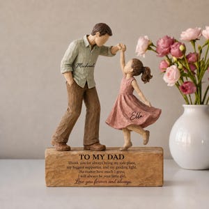 May include: A carved figurine of a person in a light green shirt labeled Michael and brown trousers, holding hands with a child in a pink dress labeled Ella. They stand on a rectangular base with the text TO MY DAD and a sentimental message.