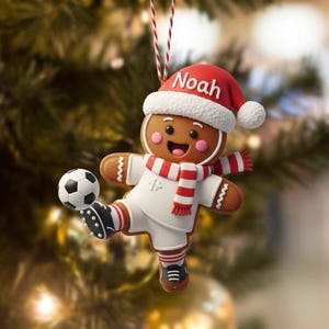 Puede incluir: Adorno navideño de hombre de jengibre con uniforme de fútbol, pateando un balón. El adorno tiene un gorro de Papá Noel rojo con el nombre "Noah", una bufanda a rayas rojas y blancas y botas de fútbol negras. El adorno cuelga de una cuerda roja y blanca.