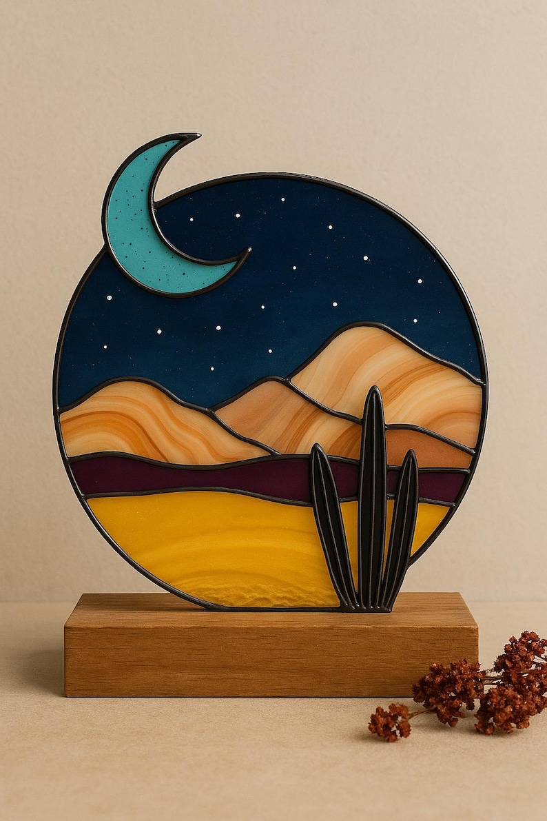 Pode incluir: Uma obra de arte em vitral com uma cena des&eacute;rtica, incluindo uma lua crescente, estrelas, montanhas e um cacto. A pe&ccedil;a circular est&aacute; montada numa base de madeira, apresentando uma paisagem noturna com tons quentes e frios.