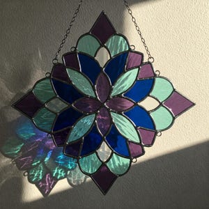 May include: A stained glass window hanging with a chain. The window is a square shape with a geometric design of purple, blue, and green glass pieces.