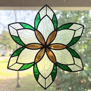 May include: A stained glass flower with six petals. The petals are a mix of clear, green, and amber glass, outlined with black. The flower is mounted in front of a window, with a blurred outdoor background visible.