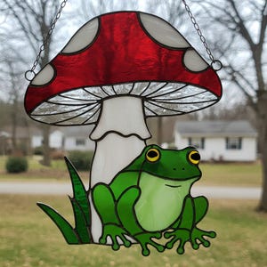 May include: Stained glass mushroom and frog suncatcher. The mushroom has a red cap with white spots and a white stem. A green frog with yellow eyes sits below the mushroom. The suncatcher hangs from a chain.