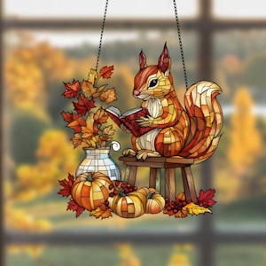 Puede incluir: Una ventana colgante de vitral representa una ardilla sentada en un taburete leyendo un libro. La ardilla está rodeada de hojas de otoño, calabazas y bayas.