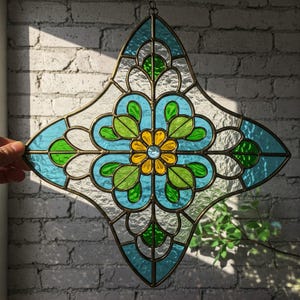 May include: Stained glass window art in a diamond shape. The design features a central yellow flower with a blue gem, surrounded by blue, green, and clear glass panels. The piece is framed with a dark metal border and is hanging in front of a brick wall.