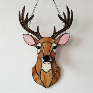 Puede incluir: Cabeza de ciervo de vidriera con cornamenta marrón, cara marrón y orejas rosadas. La cabeza de ciervo está delineada en marrón oscuro y cuelga de una cadena. La obra de arte es una pieza decorativa.