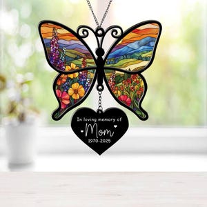 Puede incluir: Un carillón de viento de mariposa de metal negro con un diseño de vitral que presenta flores coloridas y un paisaje de puesta de sol. Un amuleto de corazón negro cuelga de la mariposa con el texto "In loving memory of Mom 1970-2025".