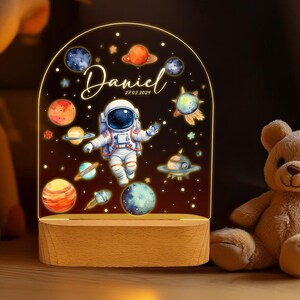 May include: A night light with a clear acrylic base and a white astronaut design. The astronaut is surrounded by planets and stars. The text "Daniel 27.02.2024" is printed on the base.