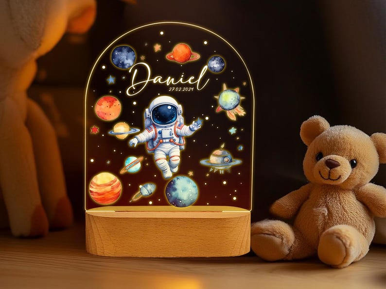 May include: A night light with a clear acrylic base and a white astronaut design. The astronaut is surrounded by planets and stars. The text "Daniel 27.02.2024" is printed on the base.