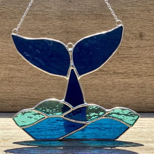 May include: Stained glass whale tail art piece. The artwork features a dark blue whale tail above a sea of blue and green waves. The piece is framed with silver metal and hangs from a silver chain.