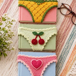 Könnte beinhalten: Drei gehäkelte Buchhüllen mit dekorativen Motiven: eine gelbe Ananas mit grünen Blättern, eine hellgrüne Hülle mit zwei roten Kirschen und grünen Blättern sowie eine rosa Hülle mit einem dunkelrosa Herz. Jede Hülle hat einen gewellten Rand.