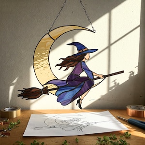 Könnte beinhalten: Buntglas-Hexe auf einem Besen, die über einen goldenen Halbmond fliegt. Die Hexe trägt ein lila Kleid und einen blauen Hut. Der Mond hängt an einer Kette. Eine Zeichnung der Hexe und des Mondes liegt auf einem Tisch.