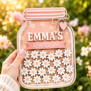 Può includere: Un barattolo premio in legno a forma di vasetto con sfondo rosa chiaro e motivi a margherite bianche. Il testo EMMA'S REWARD JAR è scritto in lettere bianche. Un piccolo cuore in legno e un fiocco di spago sono fissati in alto a destra.