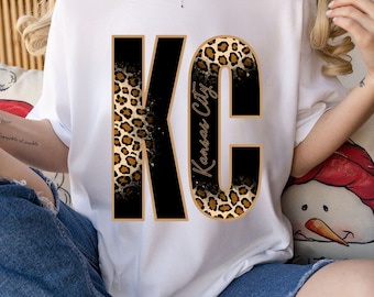 May include: White t-shirt with a black and gold "KC" graphic. The letters are filled with a leopard print pattern and the "C" has the words "Kansas City" written vertically inside it.