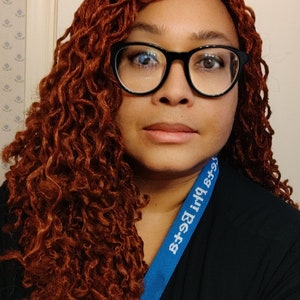 May include: A woman with long, curly auburn hair wearing black-framed glasses and a black shirt with a blue lanyard that says "8+9=17".