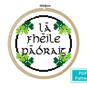 May include: A cross-stitch pattern featuring the Irish phrase "Lá fhéile Pádraig" surrounded by green shamrocks in a wooden embroidery hoop. The pattern is available as a PDF download.