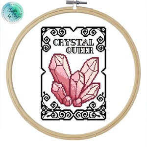 May include: A black and white cross stitch pattern with a pink crystal cluster in the center. The text "CRYSTAL QUEER" is above the crystal cluster.