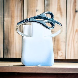 May include: A silver ring shaped like a pair of scissors. The ring is displayed on a white stand.