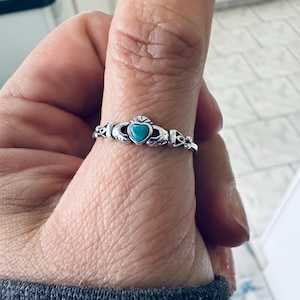 Puede incluir: Un anillo Claddagh plateado con una piedra turquesa en forma de corazón. El anillo presenta manos sosteniendo el corazón, con una corona encima. El anillo se lleva en un dedo.